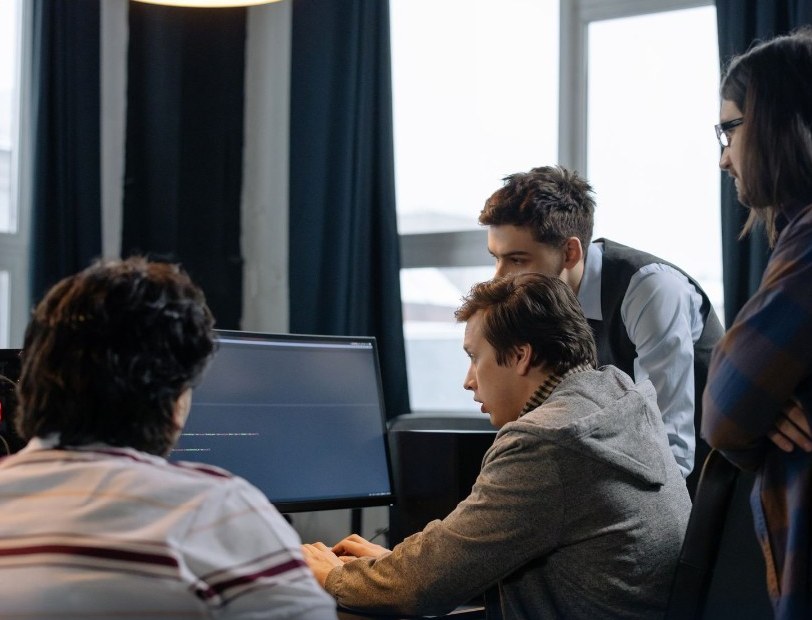 Tre ragazzi davanti a un monitor, uno scrive codice e gli altri due seguono